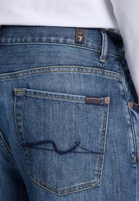 Jeans da uomo in denim blu, con una tasca posteriore caratterizzata da un design ricamato curvo, cuciture dorate e un piccolo etichetta del marchio vicino alla vita.