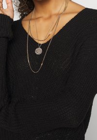 Maglione nero a maglia con una vestibilità ampia, caratterizzato da un collo a V e maniche lunghe. Multiple collane dorate con un pendente decorato.
