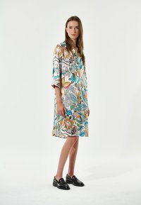Blumenmuster-Kleid mit knielangem Saum, bunten Akzenten, Knopfleiste und Drei-Viertel-Ärmeln. Kombiniert mit schwarzen Slippern.