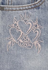 Tejido de mezclilla con un lavado azul claro que presenta corazones y hojas bordados en rosa, junto con la palabra "AMOR" en un diseño gráfico.
