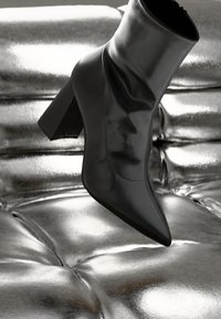 Black ankle boot with a pointed toe, made of smooth material, featuring a chunky heel and gathered design, placed on a metallic silver surface.