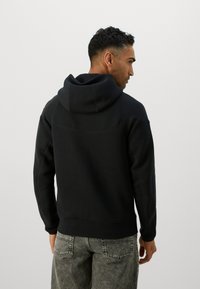 Sort hættetrøje med en tekstureret, blød stof. Har en kengurulomme og ribbede manchetter. Bæres med lysegrå slidte jeans.