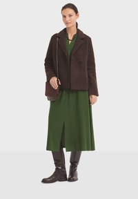 Žena oblečená v tmavo hnedej dvojradovej bunde, pod ktorou má zelené midi šaty, hnedé čižmy po kolená a nesie hnedú crossbody tašku.