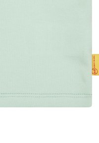 Maglietta in cotone verde menta chiaro con una texture liscia, dotata di un'etichetta gialla sul lato. L'orlo è cucito con filo abbinato.