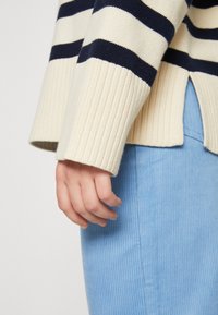 Knit striped sweater in cream with navy stripes, featuring ribbed cuffs and a side slit, paired with a light blue corduroy skirt.