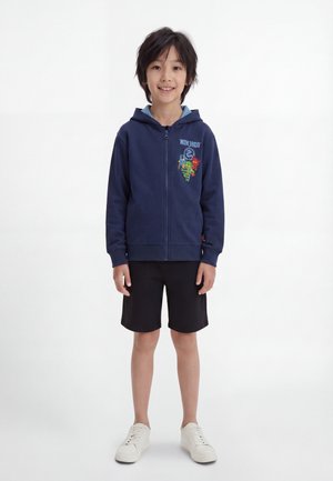 Pojke står och bär en marinblå zip-up hoodie med Ninjago-grafik, svarta shorts och vita sneakers mot vit bakgrund.