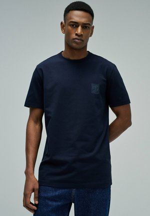 Joven de pie con una mano detrás de la espalda, vistiendo una camiseta azul marino lisa y jeans azules contra un fondo neutro.