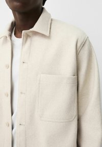 Giacca camicia in lana beige con collo, chiusura a bottoni e tasca sul petto con design piatto e texture morbida.