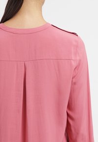 Femme aux cheveux foncés attachés, portant une chemise rose à manches longues avec une couture d'empiècement et un détail de pli au dos sur fond blanc.