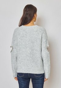 Pull en maille gris clair avec une coupe décontractée, orné d'accents argentés décoratifs sur les manches et d'un ourlet côtelé. Porté avec un jean foncé.