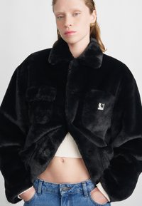 Chaqueta de pelo sintético negro con cuello, dos bolsillos delanteros y un parche de logo. Llevada encima de una camiseta de canalé color crema y jeans azules. Textura suave.