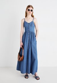 Denim maksikjole med justerbare stropper, knappestolpe foran, lommer og en flowy silhuett. Tilbehøret inkluderer en brun håndveske og solbriller.