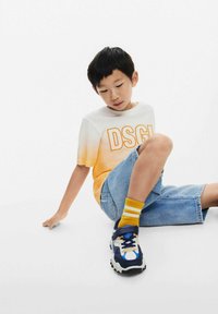La maglietta presenta un design ombre che va dal bianco all'arancione con "DSGL" in lettere grasse. Indossata con pantaloni corti in denim blu e calze gialle a righe.