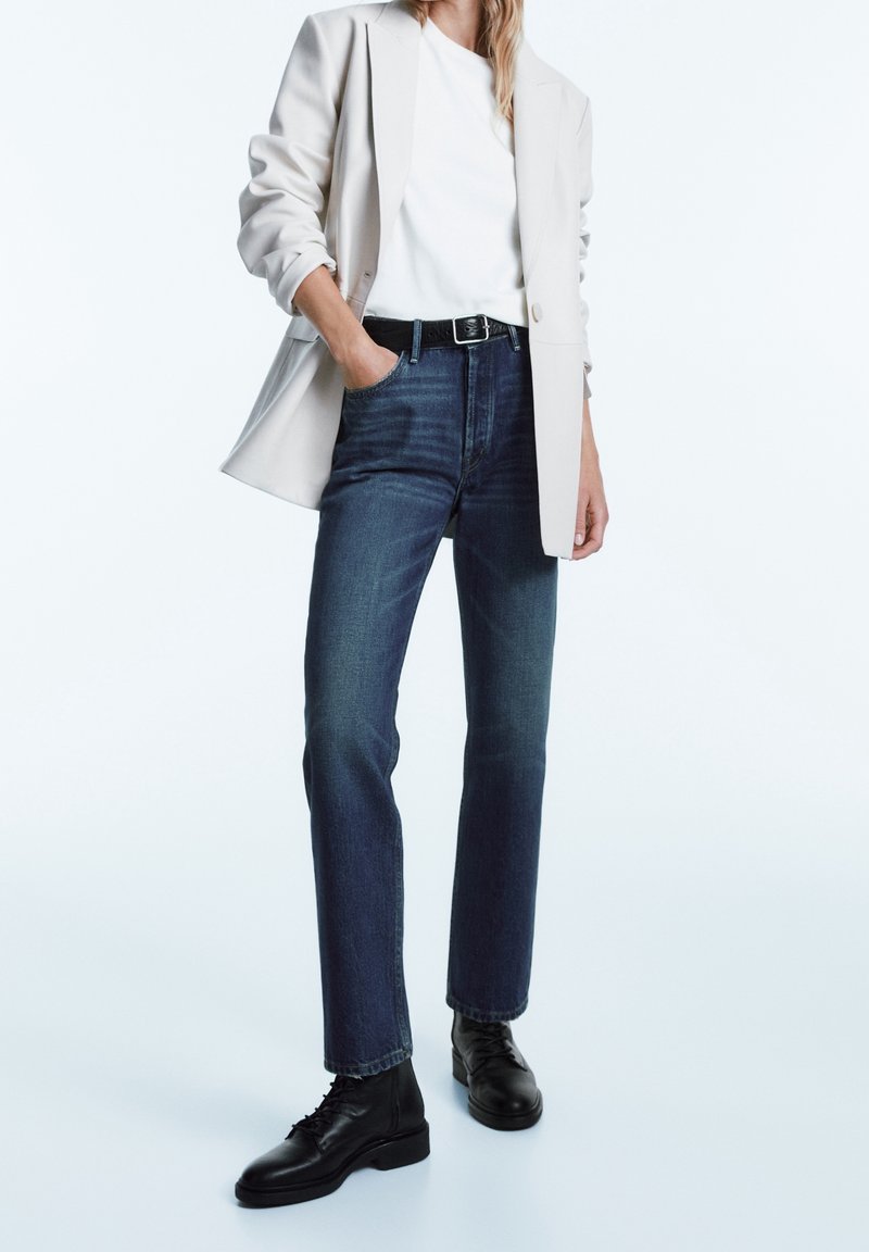 Jeans i mörkblått denim, rak passform, kombinerat med en vit t-shirt och en beige blazer. Svarta snörstövlar fulländar outfiten.