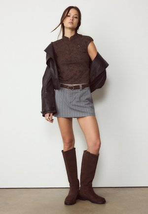 Žena stojaca opretá o bielu stenu, oblečená v bezrukávovom hnedom topu, sivej minisukni s pásikmi, tmavohnedom saku zloženom z ramien a hnedých čižmách po kolená.