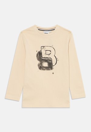 Chemise beige à manches longues en coton doux avec une lettre "B" stylisée imprimée en noir et des accents de pulvérisation subtils à l'avant.