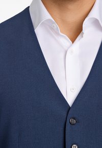 Marinblå väst med en slät yta, som har en djup V-ringning, knappstängning och är överlagd över en vit skjorta med spetskrage.