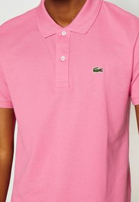 Polo rose en coton texturé, avec une patte de boutonnage à trois boutons et un petit logo crocodile vert brodé sur la poitrine.