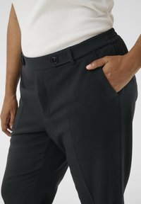 Pantalons noirs ajustés en tissu lisse, avec une taille plate, une ceinture élastique et des poches latérales inclinées. Fermeture classique à bouton.