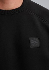 Sudadera negra con cuello acanalado y tejido texturizado. Una pequeña etiqueta negra con "PME Flying Legend" está cosida en el pecho.