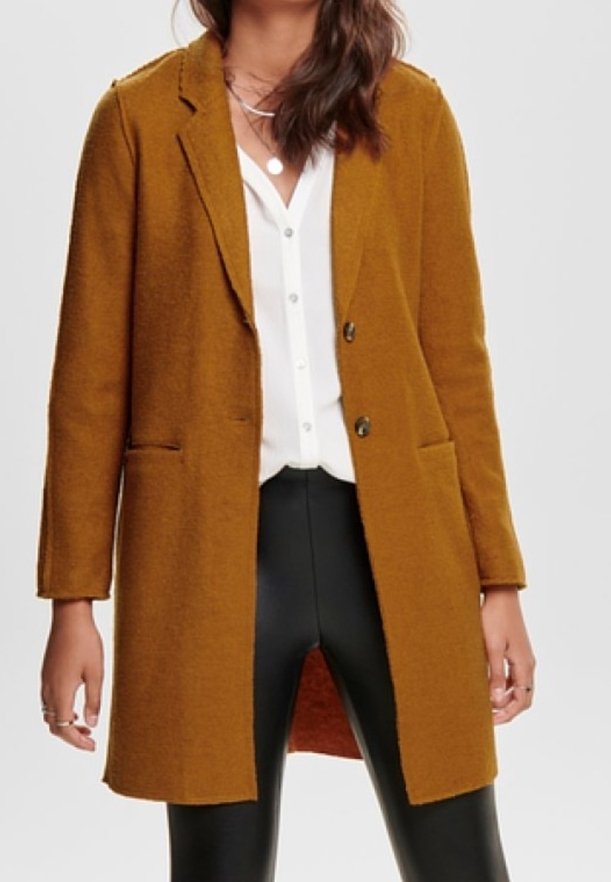 Femme portant un manteau boutonné jaune moutarde par-dessus un chemisier blanc et un pantalon en cuir noir, avec des colliers argentés superposés.