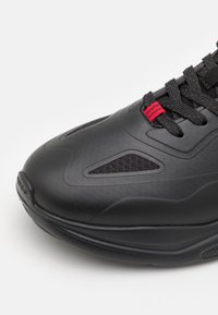 Baskets noires vue de face avec une tige lisse et mate, des empiècements en mesh respirant, des lacets noirs et un petit détail en boucle de lacet rouge.