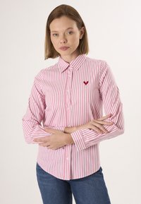 Chemise boutonnée à rayures rose et blanche avec un col, arborant un logo en forme de cœur sur la poitrine gauche et des manches longues avec des poignets boutonnés.