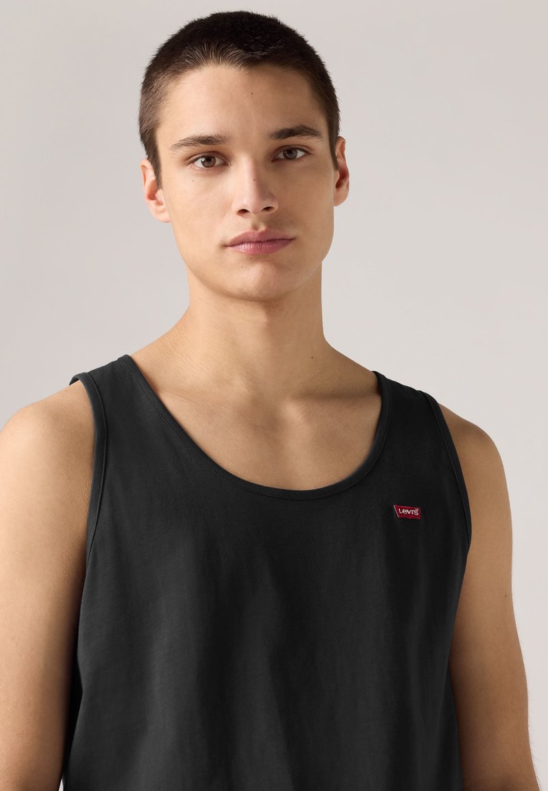 Junger Mann mit kurzen Haaren, der ein schwarzes Levi's Tanktop trägt und vor einem einfarbigen hellen Hintergrund steht.