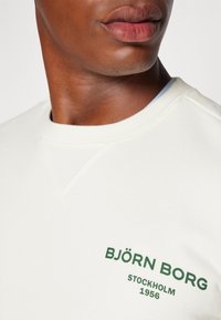 Felpa bianca con collo rotondo, caratterizzata da un ricamo verde con la scritta "BJÖRN BORG STOCKHOLM 1956" sul petto. Tessuto morbido, vestibilità casual.
