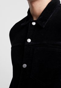 Svart manchester-skjorta med knäppning framtill, krage och bröstficka. Diskret struktur med metalliska tryckknappar framtill.