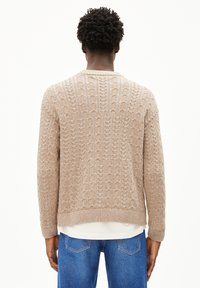 Pull en tricot beige à mailles torsadées avec une coupe décontractée, des poignets et un ourlet côtelés, et un motif texturé, porté sur une chemise blanche et associé à un jean bleu.