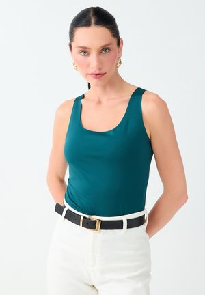 Femme aux cheveux foncés attachés en queue de cheval, portant un haut sans manches bleu sarcelle, un pantalon blanc, une ceinture noire et des boucles d’oreilles créoles dorées sur fond uni.