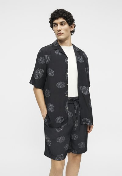 Νεαρός άνδρας που φοράει μαύρο πουκάμισο με κουμπιά και αντίστοιχο σορτς με λευκά φύλλα πάνω από λευκό T-shirt, στέκεται με τα χέρια στις τσέπες.