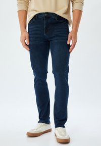 Jeans de mezclilla azul oscuro con corte slim, con sutiles rayas horizontales en el frente, combinados con zapatillas color crema y un suéter beige.