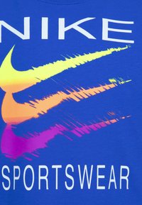 Modrá tričko s veľkým, viacfarebným logom Nike swoosh a bielym nápisom "SPORTSWEAR." Materiál sa zdá byť hladký a má štandardný strih.