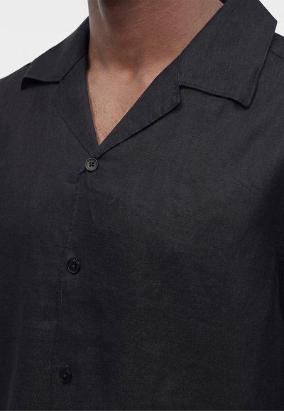 Gros plan d'une personne portant une chemise en lin noire avec un col camp et trois boutons noirs visibles sur le devant.