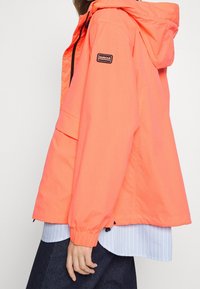 Helle, orangefarbene, leichte Jacke mit Kapuze, elastischen Bündchen und einer Fronttasche. Verfügt über ein schwarzes Logo-Patch auf dem linken Ärmel.