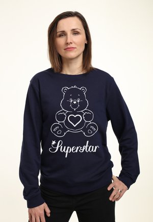 Marineblå sweatshirt lavet af blødt stof, med et hvidt bjørne grafik, der har et hjerte og teksten "Superstar" i et legende skrifttype.