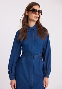 Niebieska sukienka dżinsowa z długimi rękawami, kołnierzykiem zapinanym na guziki, talią zdobioną paskiem oraz kontrastowymi szwami. Modelka nosi duże, kwadratowe okulary przeciwsłoneczne.