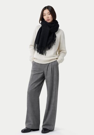 Frau trägt einen weißen Pullover, schwarzen Fransen-Schal, weite graue Hosen und schwarze Schuhe, steht mit den Händen in den Taschen.