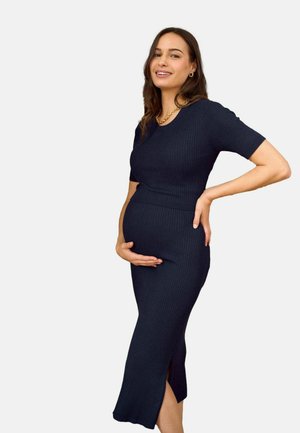 Raskaana oleva nainen istuvassa, merensinisenä ribbineulottuun midimekkoon hymyillen, toinen käsi vatsalla ja toinen lantiolla.