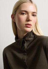 Braune gerippte Knopfleiste mit umgeschlagenem Kragen, ausgestattet mit sechs Knöpfen und einem strukturierten Stoff. Glatte Haut mit dezentem Make-up und glattem, blonden Haar.