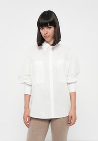 Camicia bianca con colletto, due tasche frontali e maniche ampie. Il tessuto sembra liscio e leggero. Abbinata a pantaloni beige.