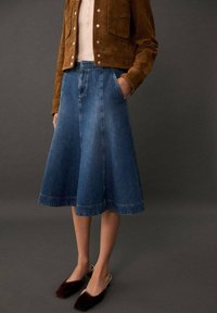 Džínová sukně s rozšířeným střihem, délka ke kolenům, s hlubokými bočními kapsami a zesvětleným vzhledem. V kombinaci s hnědou semišovou bundou a tmavými botami.