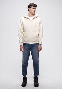 Hanorac crem cu fermoar frontal, buzunare mari și glugă căptușită, asortat cu blugi albaștri conici și cizme negre, stând pe un fundal neutru.