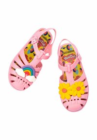Sandalias de plástico rosa con acentos de arcoíris y nubes en un pie y flores amarillas en el otro, con una plantilla de colorido patrón.