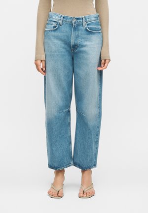 Άτομο που φοράει ανοιχτό μπλε τζιν παντελόνι με φαρδιά μπατζάκια, μπεζ ριμπ μπλούζα με μακριά μανίκια και μπεζ σανδάλια με τετράγωνη μύτη, στέκεται μπροστά σε λευκό φόντο.