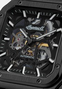 Schwarze Skelettuhr mit quadratischem Gehäuse, die aufwendige Zahnräder, goldene Akzente und leuchtende Stundenmarker präsentiert. Verfügt über Automatikwerk.