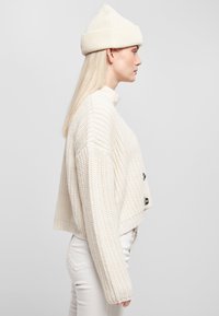 Avskuren, krämfärgad cardigan i ribbad stickning med hög krage och svarta knappar. Matchas med ljusa, slim-fit byxor och en matchande mössa.