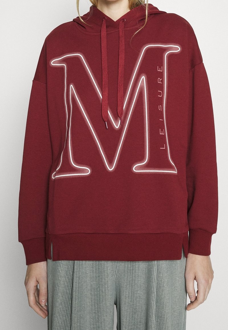 Personne portant un sweat à capuche bordeaux avec une grande lettre "M" blanche en contour et le mot "Leisure" verticalement sur le devant.